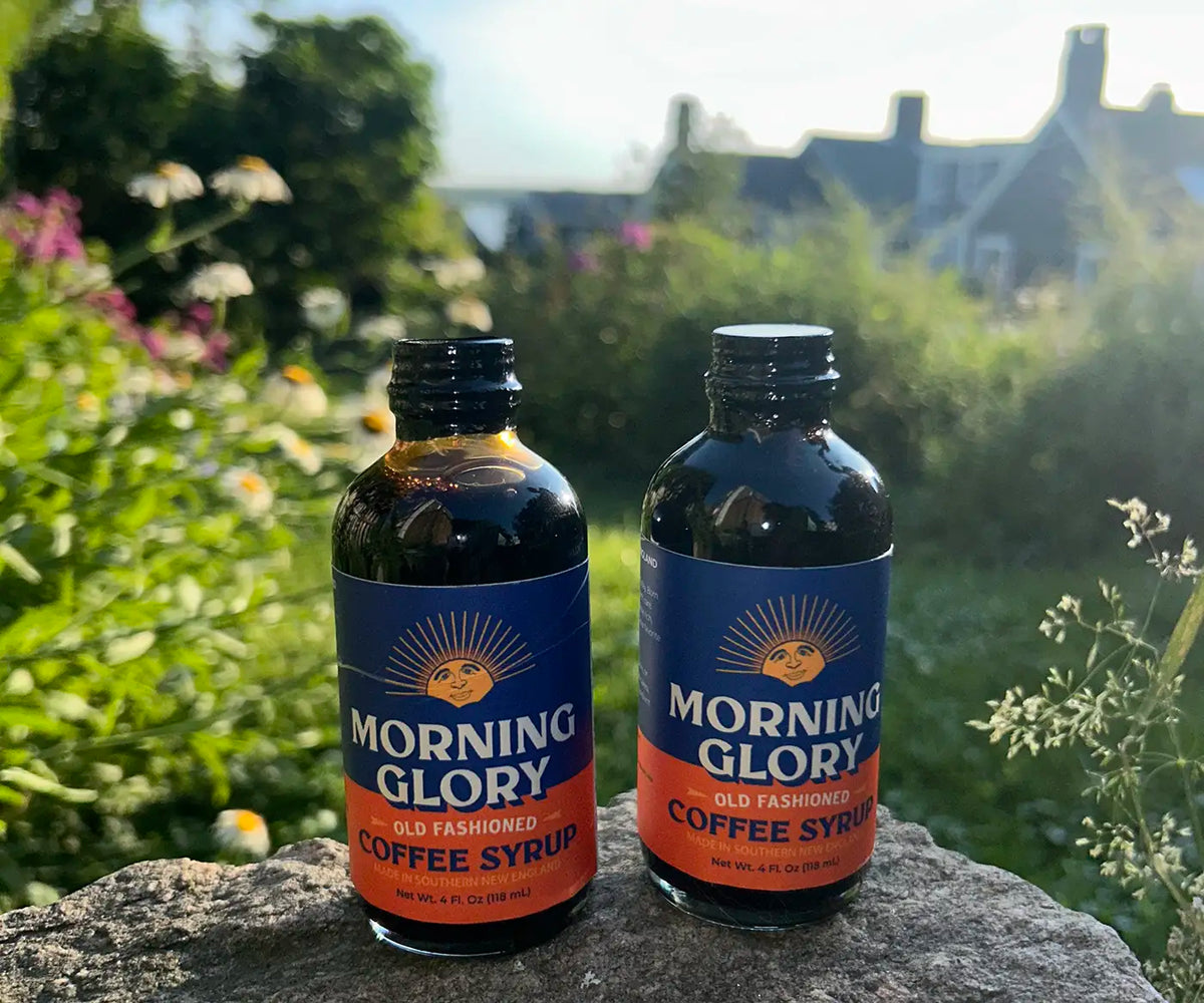 Two bottles of Morning Glory Old Fashioned Coffee Syrup standing on a stone surface outdoors, with a blurred garden and house in the background.