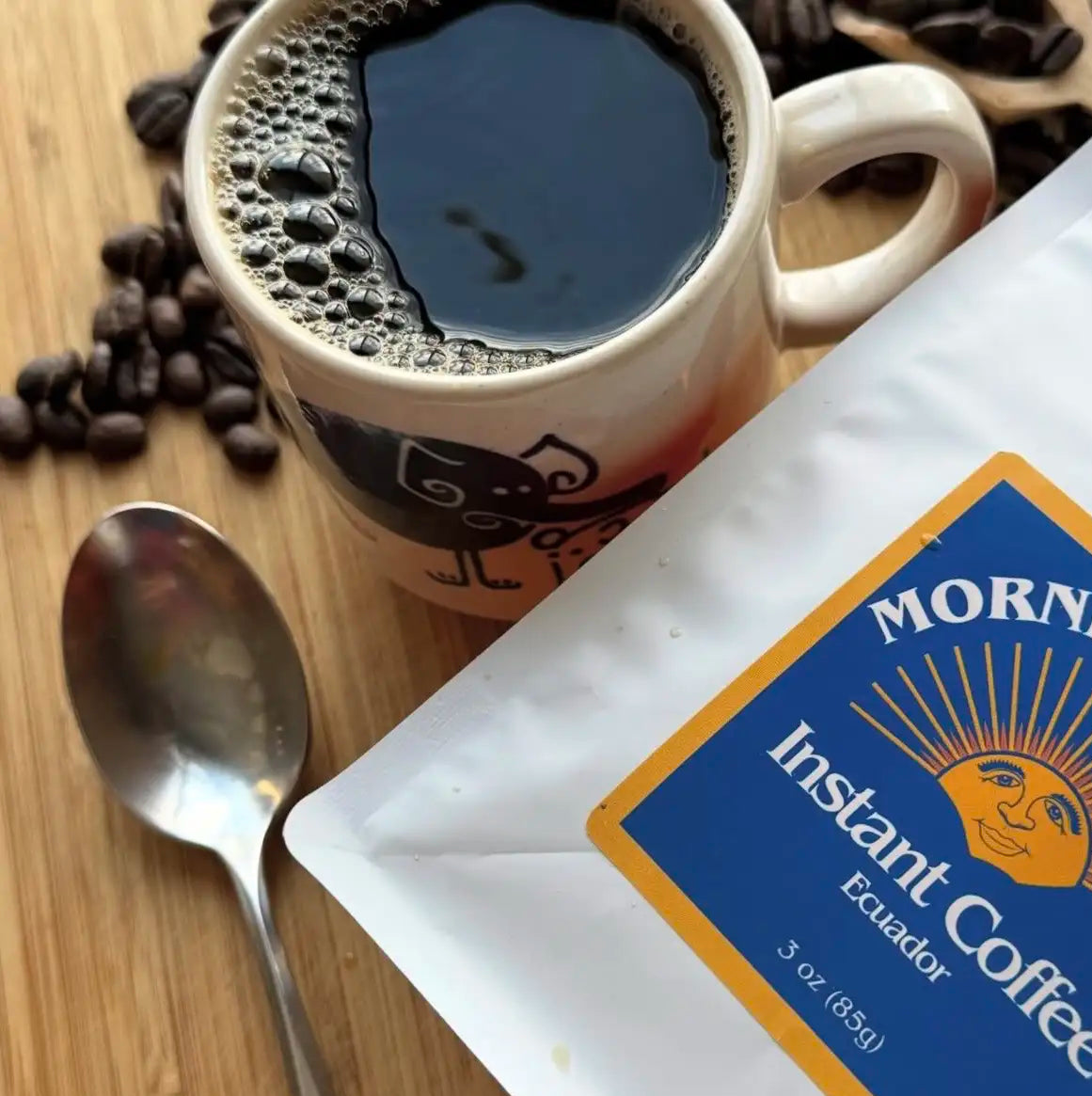 A white bag of Morning Instant Coffee Ecuador with a blue and yellow label, shown next to a mug of black coffee, scattered coffee beans, and a spoon on a wooden surface.