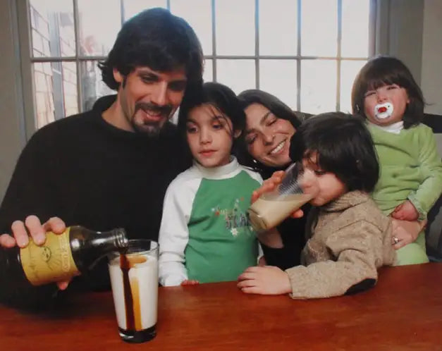 A family portrait showing a man, a woman, and three children gathered around a wooden table. The man on the left is pouring a dark brown liquid from a bottle into a glass of milk, while the woman and children drink from glasses.