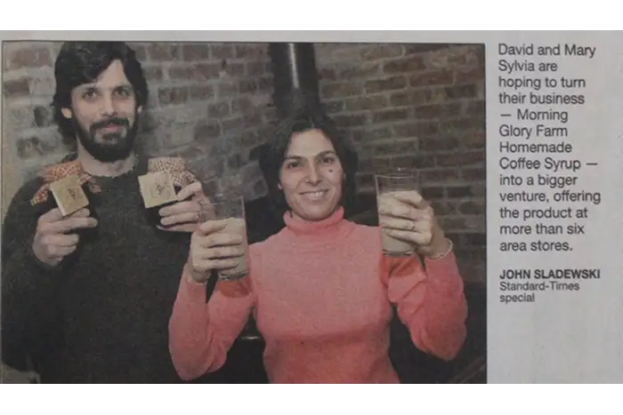 A newspaper-style photo with a brick background shows the owners holding small boxes and glasses of coffee syrup. A text block on the right reads about David and Mary Sylvia hoping to turn their business into a bigger venture, with the headline Morning Glory Farm Homemade Coffee Syrup and a byline for John Sladewski, Standard-Times special.